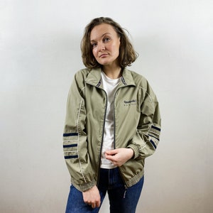 Könnte beinhalten: Eine hellgrüne Windbreaker-Jacke mit einem weißen Reißverschluss und schwarzen und weißen Streifen an den Ärmeln. Die Jacke hat das Reebok Classics Logo auf der Vorderseite.