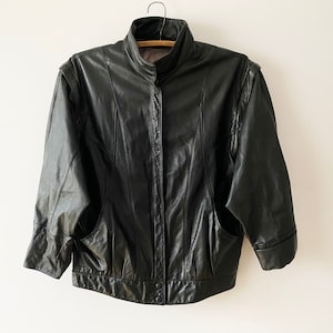 Puede incluir: Una chaqueta de cuero negra con cuello alto y cierre de cremallera. La chaqueta tiene una longitud ligeramente recortada y presenta dos bolsillos en la parte delantera.