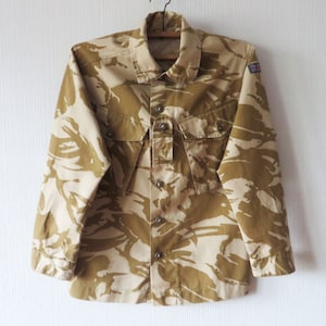 Può includere: Una camicia a maniche lunghe con motivo mimetico beige e marrone, con chiusura a bottoni sul davanti e una toppa con la bandiera dell'Unione Jack sulla manica destra.