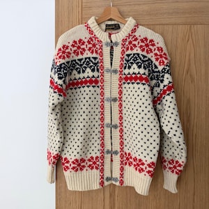 May include: A cream-colored knit cardigan featuring a red and navy blue Nordic-inspired pattern across the chest and shoulders. The sweater has a ribbed collar, cuffs, and hem, and is fastened down the front with five decorative silver-toned metal clasps.