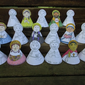 May include: A collection of 15 paper cone figures of Catholic saints, each with a different image and name. The figures are all white with colorful details and are arranged in three rows on a wooden surface.