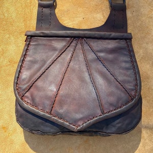 May include: A brown leather satchel with a flap closure and a long strap. The bag has a decorative stitching pattern and a slightly curved shape.