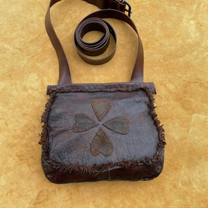 May include: A brown leather satchel with a fringe trim and a four-leaf clover design. The bag has a long adjustable strap.