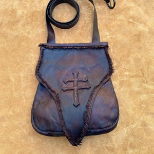 May include: A brown leather satchel with a fringe trim and a cross design. The bag has a long adjustable strap.