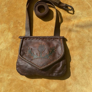 May include: A brown leather crossbody bag with a flap closure and a long strap. The bag features a decorative thistle design on the front. The strap is adjustable and has a buckle. The bag is set against a yellow background.