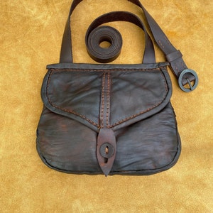 May include: A dark brown leather satchel with a flap closure and a long strap. The bag features visible stitching in a lighter brown thread. A separate, rolled-up leather strap is also present, along with a metal buckle.