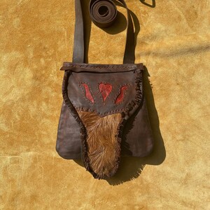 May include: A brown leather bag with a long strap and a fringe detail. The bag has a red and brown design on the front and a patch of brown fur.