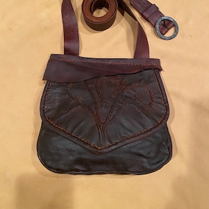 May include: A brown leather bag with a flap and a decorative design. The bag has a long strap with a metal buckle.