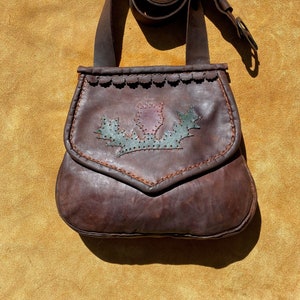 May include: A brown leather shoulder bag with a flap closure. The bag features a decorative stitched design of a flower and leaves in shades of purple and green. The strap is rolled up and attached to the bag.