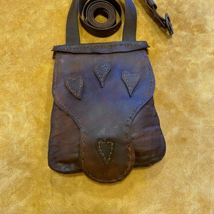 May include: A dark brown leather crossbody bag with three heart-shaped cutouts and a long strap. The bag has a flap closure and is made of textured leather. The strap is adjustable and the bag appears to be handmade.