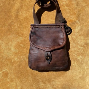 May include: A small, brown leather shoulder bag with a flap closure and a long, adjustable strap. The bag features a button closure and decorative stitching along the top edge. The leather has a distressed appearance.
