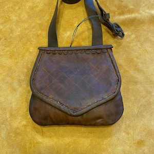 May include: A brown leather shoulder bag with a flap closure and a long strap. The bag features a diamond pattern on the front and decorative stitching along the top edge. The strap is adjustable and has a buckle.