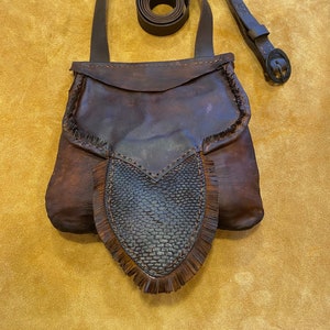 May include: A handcrafted brown leather shoulder bag with a textured front flap and fringe detailing. The bag features a long, adjustable strap and a matching belt. The leather has a distressed appearance, giving it a rustic aesthetic.