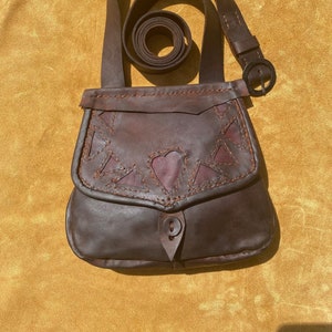 May include: A brown leather satchel bag with a flap closure and a decorative heart design. The bag has a long strap and a buckle closure.