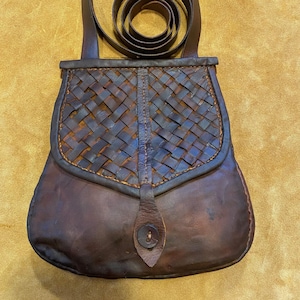 Puede incluir: Una bandolera de cuero marrón con una solapa tejida y una correa larga. La bolsa tiene un cierre de botón único.