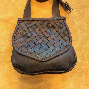 May include: A brown leather bag with a woven design and a long strap. The bag has a flap with a pointed bottom.