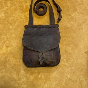 May include: A dark brown leather shoulder bag with a flap closure and a long strap. The bag has a textured surface with a stitched design and a decorative button closure. The strap is rolled up and is approximately 40 inches long.