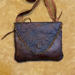 May include: A brown leather crossbody bag with a frayed edge and a single button closure. The bag has a long strap made of woven burlap with red stripes.