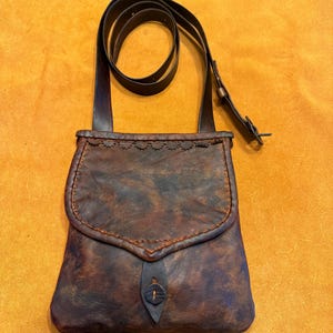 May include: A dark brown leather crossbody bag with a mottled finish, featuring a fold-over flap with a decorative scalloped edge, visible orange stitching, and a single leather button closure. It includes a long, adjustable leather shoulder strap.