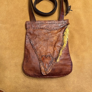 May include: A handcrafted brown leather shoulder bag with a heart-shaped accent and yellow fur trim. The bag features a long, adjustable strap and a buckle closure. The leather has a textured appearance.