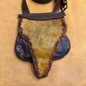 May include: A handmade leather satchel with a fur front and a long, adjustable strap. The bag features a dark brown leather body with a lighter brown fur panel and is stitched with a contrasting thread. The strap is also dark brown and has a metal buckle.