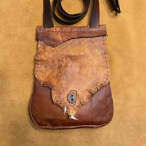 May include: Handmade leather shoulder bag in shades of brown. The bag features a flap closure with a button and a long, adjustable strap. The leather has a distressed appearance, with visible stitching and a unique texture.