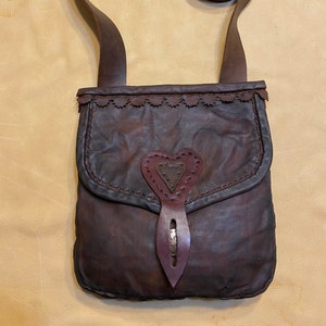 May include: A brown leather satchel with a heart-shaped flap and a decorative buckle. The bag has a long strap that can be worn over the shoulder.