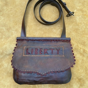 May include: A brown leather bag with a red embroidered border and the word "LIBERTY" stitched in red on the front. The bag has a long brown leather strap.