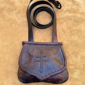 May include: A brown leather bag with a cross design and a long strap. The bag is closed with a flap and has a stitched edge.