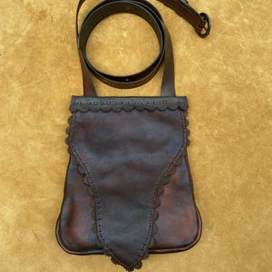 May include: A brown leather bag with a decorative scalloped edge and a long strap. The bag has a pointed flap and is made of a dark brown leather.