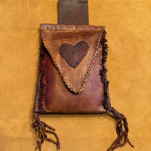 May include: Handmade brown leather pouch with a heart-shaped appliqué. The pouch features a flap closure and decorative fringe. The leather has a distressed look, with visible stitching and a dark brown heart. The pouch has a loop on the back.