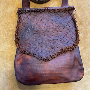 May include: A handcrafted, burgundy leather crossbody bag. The flap features a quilted diamond pattern with small perforations and a fringed edge. The bag has a long strap and a rich, textured appearance.