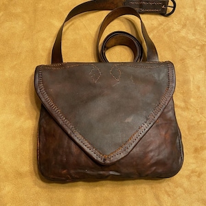 May include: A brown leather satchel with a flap closure and a shoulder strap. The bag features decorative stitching along the edges and a belt with a metal buckle. The leather has a distressed appearance.