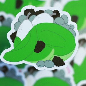 May include: A green leaf-shaped sticker with a white pear and black berries inside. The sticker is designed to look like a pear being wrapped in a leaf.