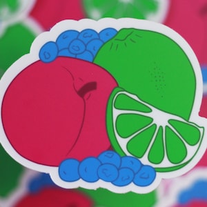 May include: A sticker featuring a pink peach, a green lime, and blue blueberries. The lime is cut in half, showing the inside. The sticker is on a white background.