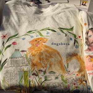 May include: White t-shirts with colorful illustrations. One shirt features a golden dog with the word "dogsboro" and a building. Another shirt displays portraits and a butterfly. Floral and architectural elements are also present.
