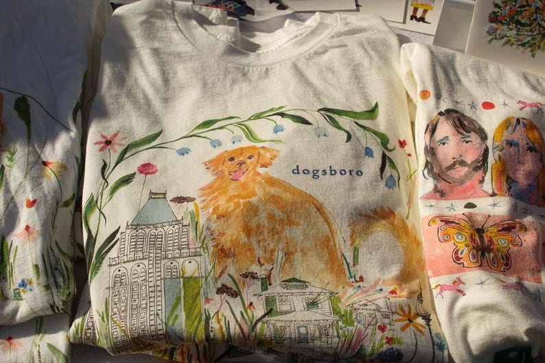 May include: White t-shirts with colorful illustrations. One shirt features a golden dog with the word "dogsboro" and a building. Another shirt displays portraits and a butterfly. Floral and architectural elements are also present.