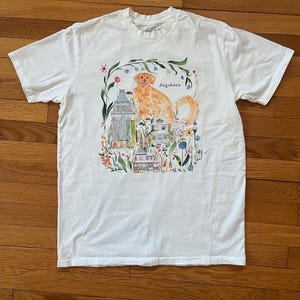 May include: White t-shirt featuring a watercolor illustration of a golden retriever dog with the word "dogsboro" above it. The design includes buildings and floral elements, creating a whimsical scene.