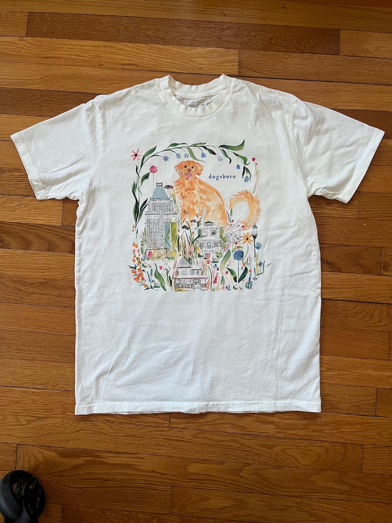 May include: White t-shirt featuring a watercolor illustration of a golden retriever dog with the word "dogsboro" above it. The design includes buildings and floral elements, creating a whimsical scene.