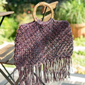 Op de afbeelding: Een donkere, meerkleurige macramé handtas met een gestructureerd knooppatroon in tinten dieppaars, blauw en zwart. De tas is voorzien van een rond, lichtgekleurd houten handvat en een lange, gefranjerde onderkant met loshangende koorden.