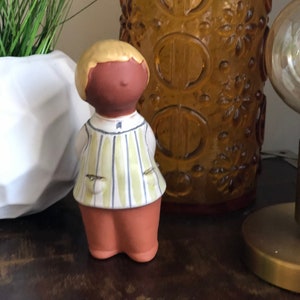 Puede incluir: Una figurita de cerámica de una persona con cabello rubio, que lleva una camisa a rayas y pantalones marrones. La figurita está de pie sobre una superficie de madera.