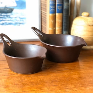 Può includere: Due salse in ceramica marrone con manici. La salsa più piccola è in primo piano e la più grande è sullo sfondo.