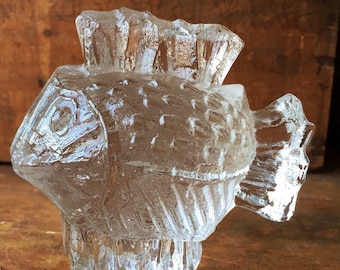 May include: A clear glass sculpture of a fish with a textured surface. The fish has a fin on its back and a tail fin.