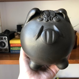 May include: A black ceramic piggy bank with a textured surface and a detailed snout. The piggy bank has a whimsical expression with large eyes and a textured mane.