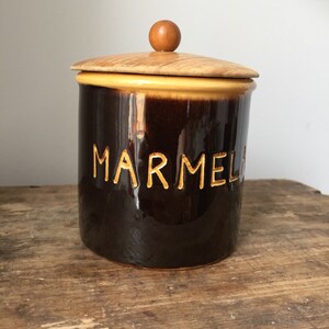 May include: A brown ceramic marmalade jar with a wooden lid and a small wooden knob. The word "MARMELADE" is carved into the jar. The jar has a glossy finish and sits on a wooden surface.