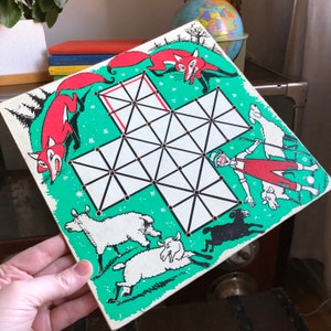 May include: A green and white game board with a grid pattern. The board features illustrations of a fox, sheep, and a person in a red and white outfit. The game board is titled "Fox and Geese".