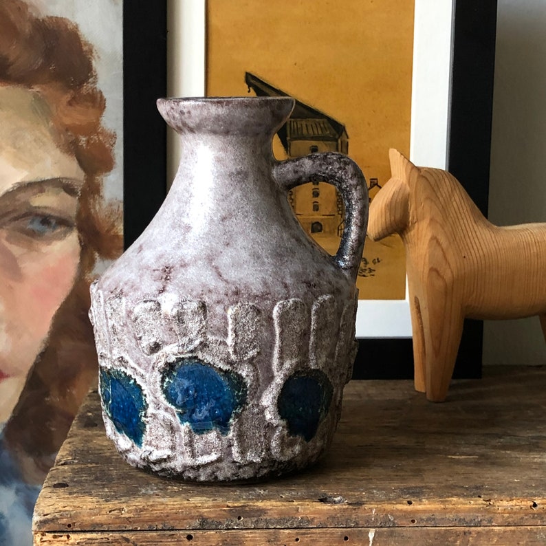 May include: A gray ceramic pitcher with blue and white accents. The pitcher has a textured surface and a wide, round base. It has a handle on the side and a spout on the front.