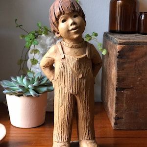 Puede incluir: Una figurita de cerámica de un niño con un mono. La figurita está pintada en tonos marrones y tiene una expresión caprichosa.