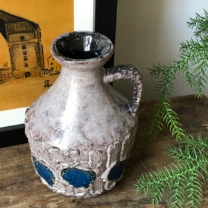 May include: A ceramic vase with a textured, gray and white glaze. The vase has a round body, a short neck, and a handle. It features blue accents around the base.