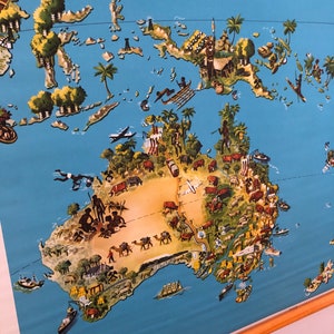 Puede incluir: Un mapa ilustrado vintage de Australia con un fondo azul y un contorno marrón del continente. El mapa presenta varios animales, personas y paisajes, incluyendo canguros, ovejas, ganado y pueblos indígenas. El mapa también incluye una pequeña ilustración de un avión volando sobre el continente.